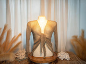 a mannequin wearing a beige knitted top with a tie detail, displayed on a wooden stand. The mannequin is positioned in front of a white curtain, and there are some decorative elements, such as a feather and a small white flower, placed around the mannequin.