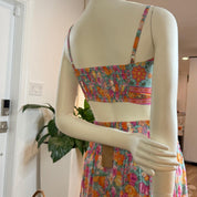 A mannequin wearing a colorful floral print dress and skirt, with a potted plant visible in the background.