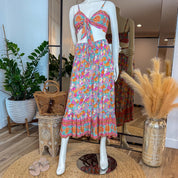 A mannequin wearing a colorful floral print dress and top, with a matching skirt, stands in a clothing store surrounded by potted plants and decorative items.