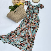 a floral print dress with a long, flowing skirt, laid out on a white background. Next to the dress is a woven straw bag, and there are yellow flowers nearby.