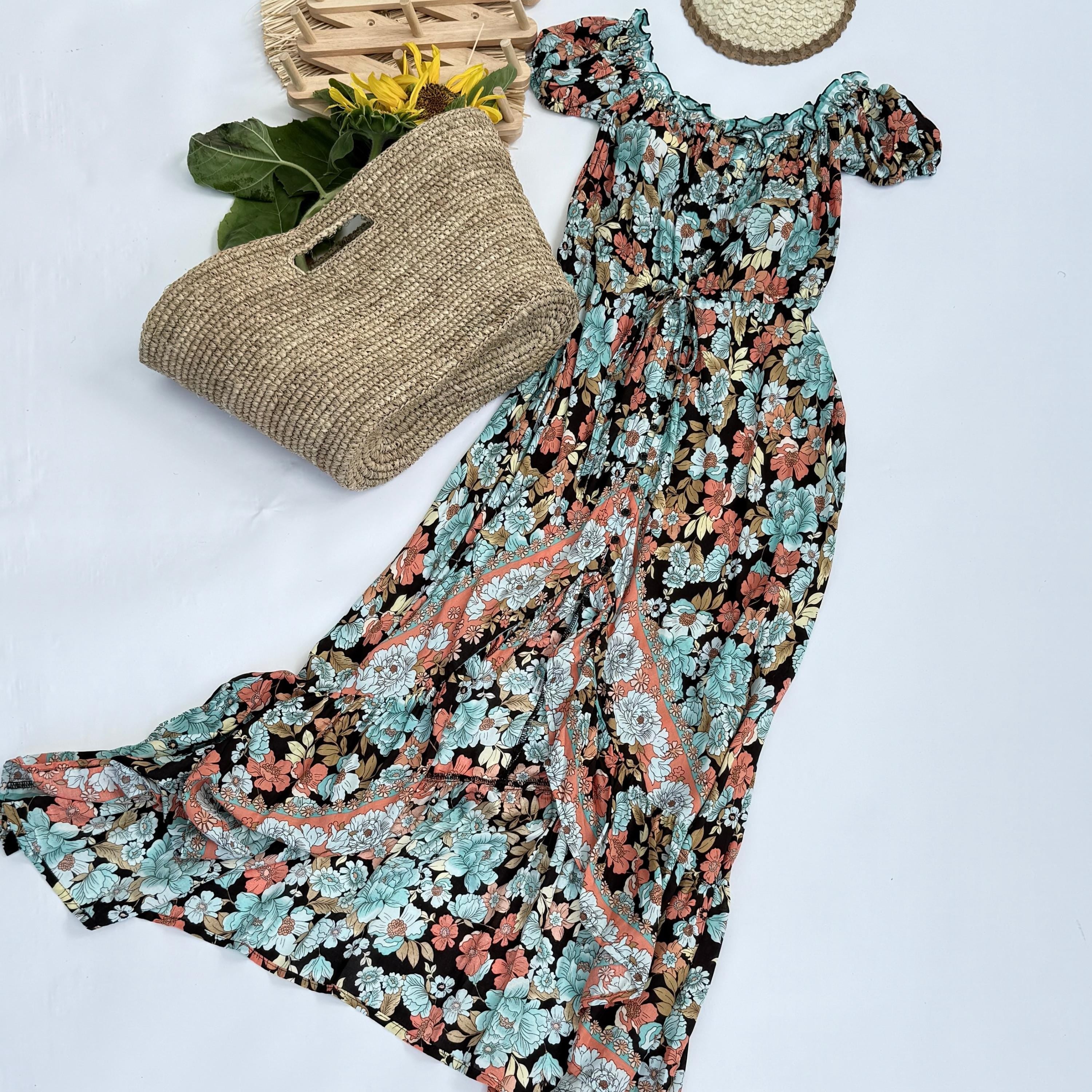 a floral print dress with a long, flowing skirt, laid out on a white background. Next to the dress is a woven straw bag, and there are yellow flowers nearby.
