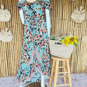 A mannequin wearing a colorful floral print dress stands next to a woven basket filled with yellow flowers, placed on a wooden stool against a bamboo wall.