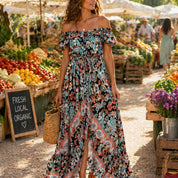 A woman in a colorful floral dress stands in front of a fruit and vegetable stand at an outdoor market.