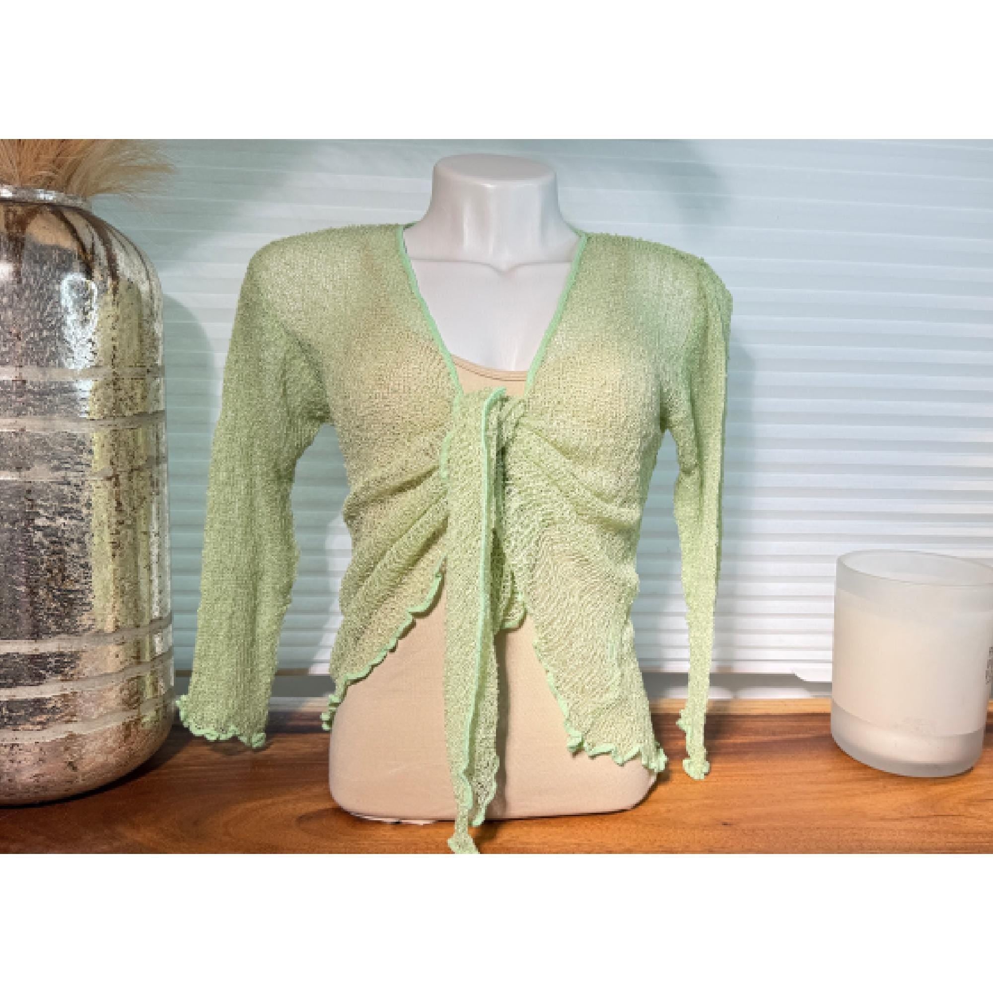 A mannequin wearing a mint green, tie-front top with long sleeves, displayed on a wooden surface next to a glass vase and a candle.