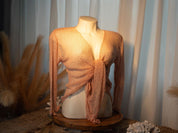 A mannequin wearing a peach-colored knitted top with long sleeves, displayed on a wooden stand against a backdrop of white curtains.