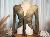 A mannequin wearing a green knitted top with a tie front, displayed on a wooden stand with a white cloth and some decorative elements in the background.