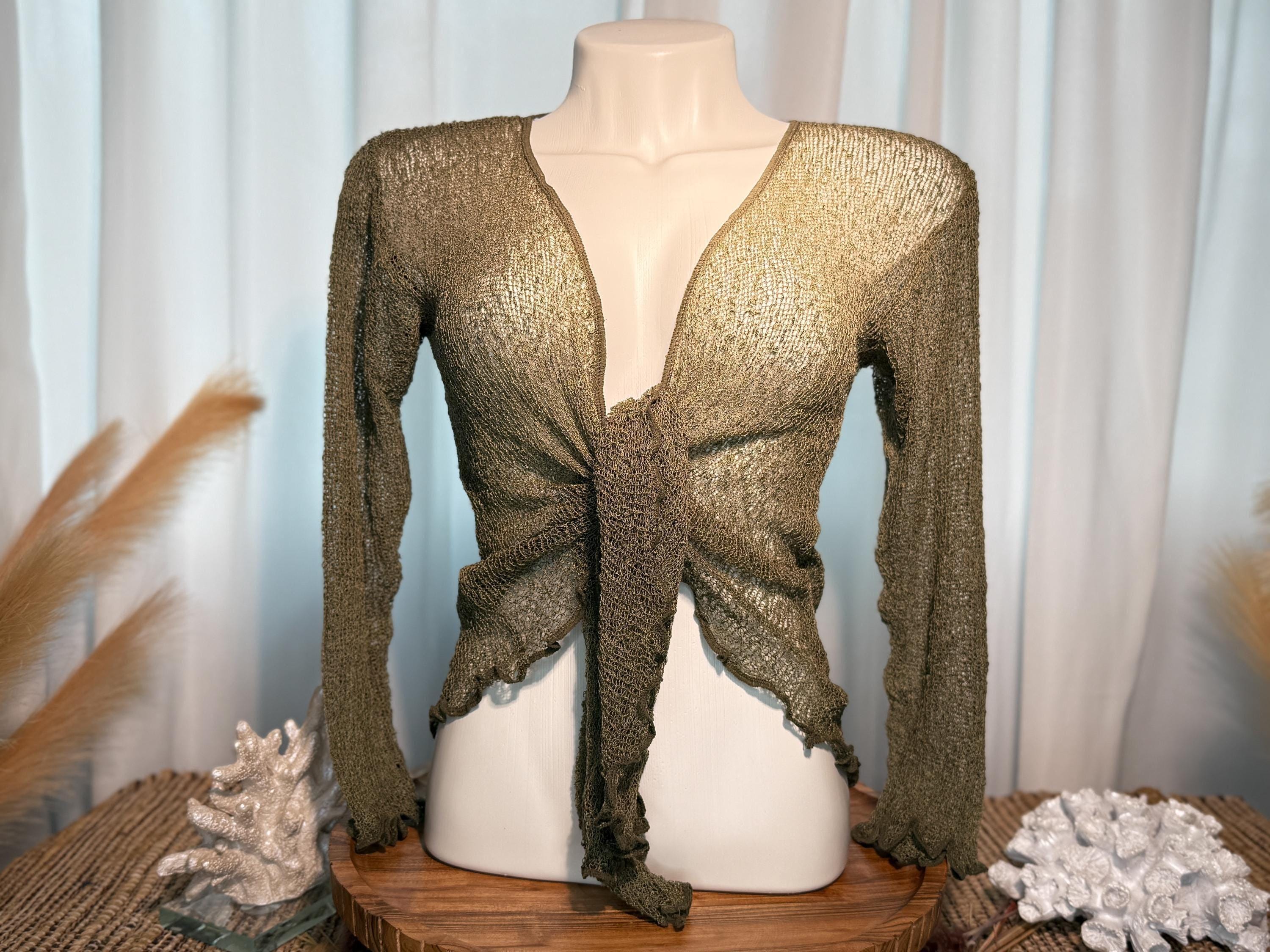 A mannequin wearing a green knitted top with a tie front, displayed on a wooden stand with a white cloth and some decorative elements in the background.