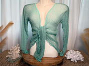 A mannequin wearing a mint green knotted top stands on a wooden stand, accompanied by a small white coral and a glass figurine.