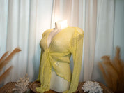 A mannequin wearing a yellow lace top with long sleeves, displayed on a wooden stand with a white curtain in the background.