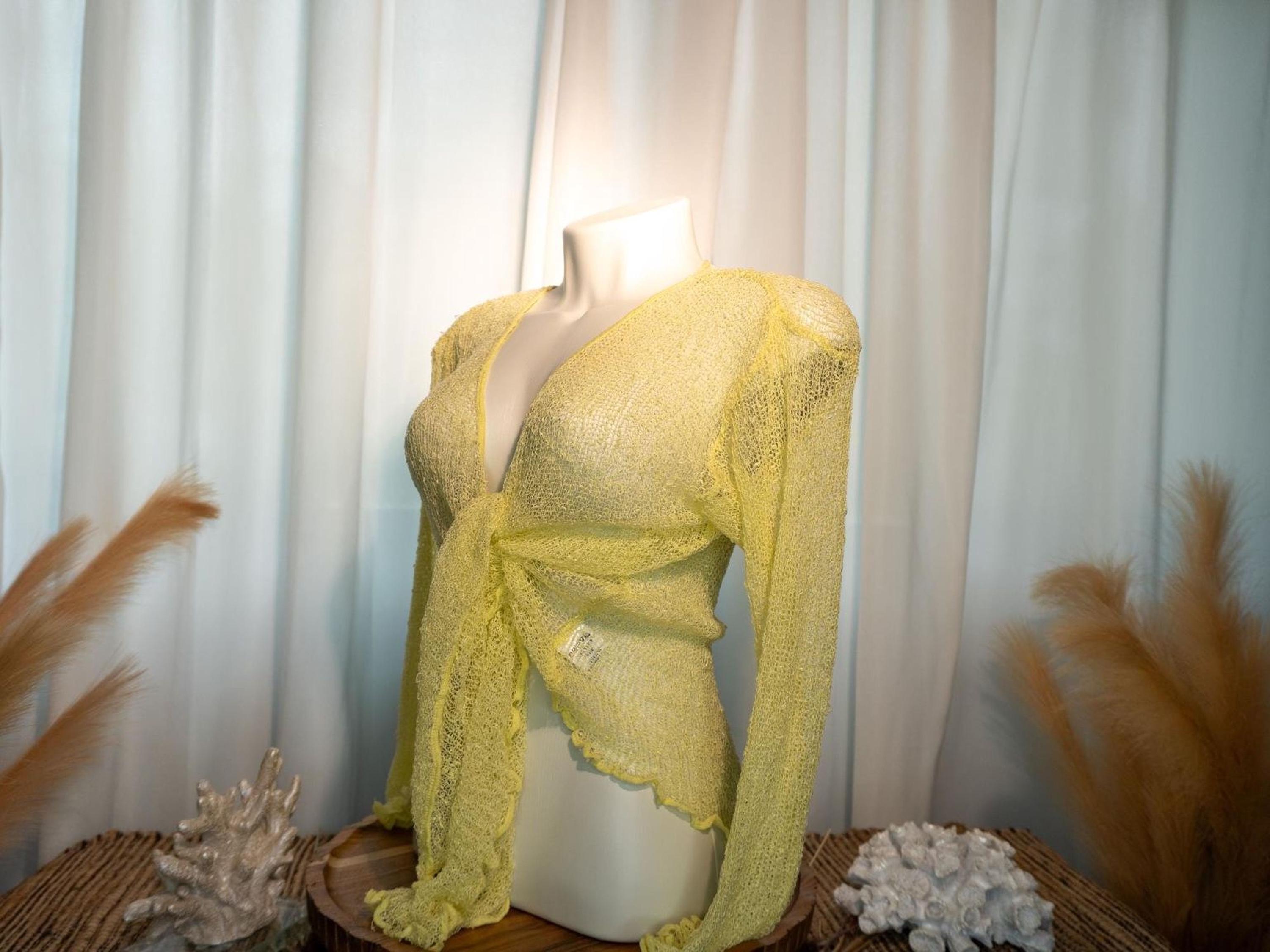 A mannequin wearing a yellow lace top with long sleeves, displayed on a wooden stand with a white curtain in the background.