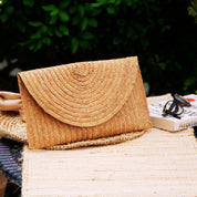 A straw purse with a curved top sits on a woven placemat, accompanied by a pair of sunglasses and a book titled "Atomium" on a table.