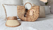 A woven straw handbag with a tag attached, a beige sun visor, and a pair of sunglasses are arranged on a white bedspread.