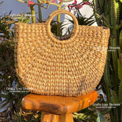 A woven straw handbag with a round handle, placed on a wooden stool in front of a cactus plant.