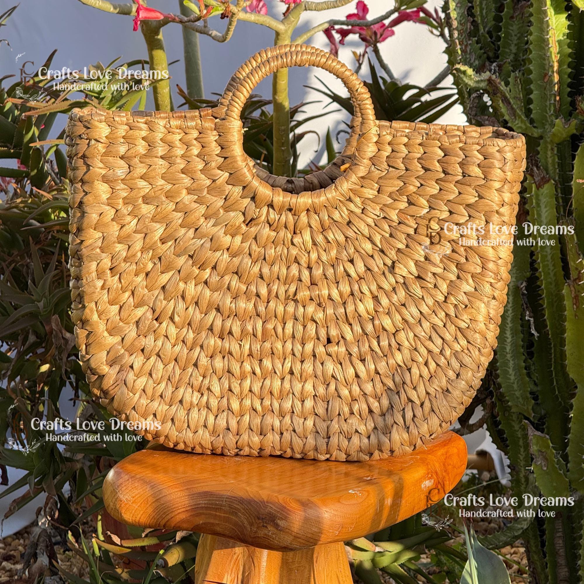 A woven straw handbag with a round handle, placed on a wooden stool in front of a cactus plant.