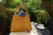 A woven straw handbag with a blue and white pattern sits on a table, accompanied by a white teacup and a book, with lush greenery and a black wrought iron chair in the background.