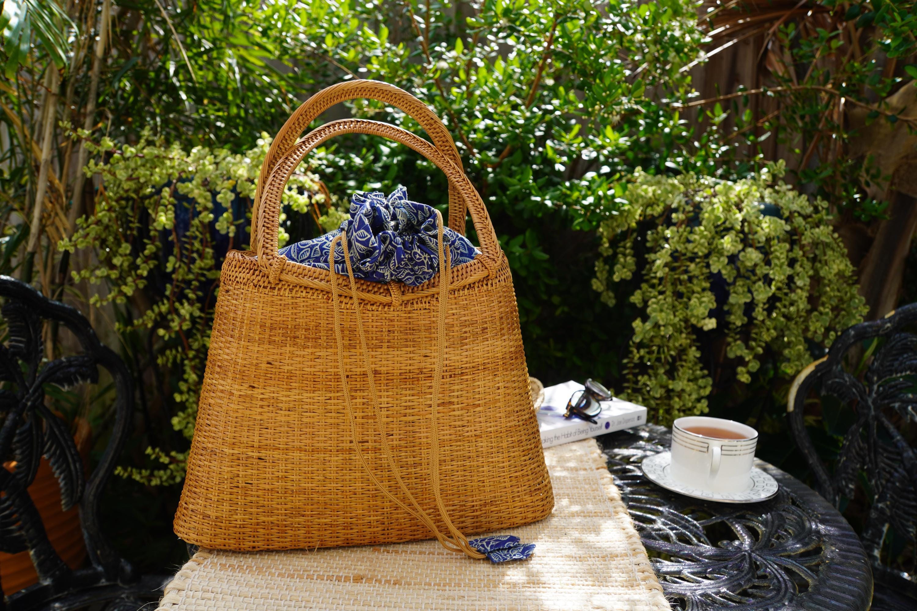 A woven straw handbag with a blue and white pattern sits on a table, accompanied by a white teacup and a book, with lush greenery and a black wrought iron chair in the background.