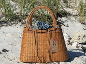 A brown woven handbag with a blue and white patterned fabric draped over the top, placed on a sandy beach with some grass and rocks in the background.