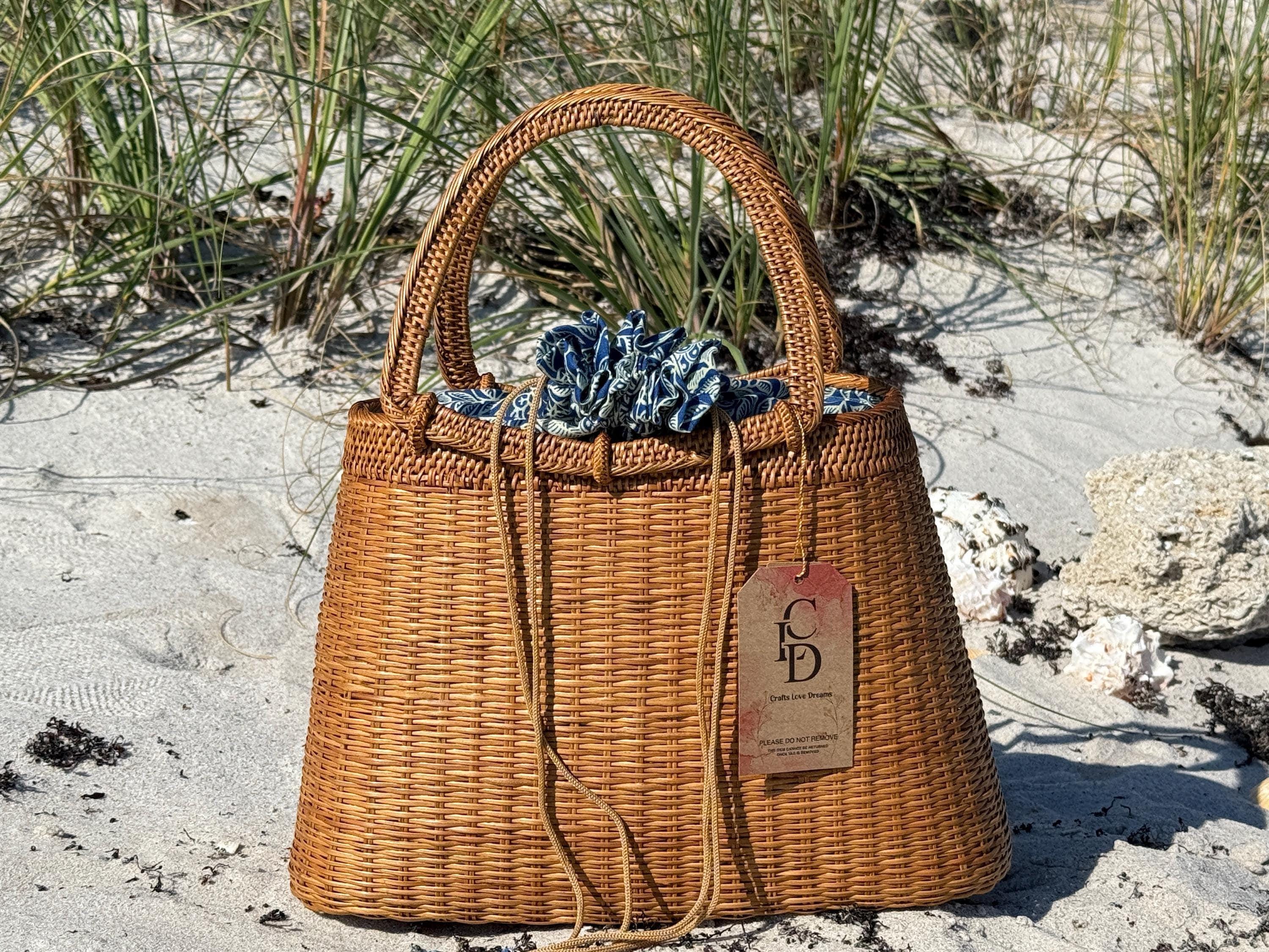 A brown woven handbag with a blue and white patterned fabric draped over the top, placed on a sandy beach with some grass and rocks in the background.