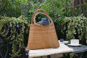 A woven straw handbag with a blue cloth tied around it is placed on a small table, with a white cup and a book nearby. The setting appears to be a garden or outdoor area with lush greenery in the background.