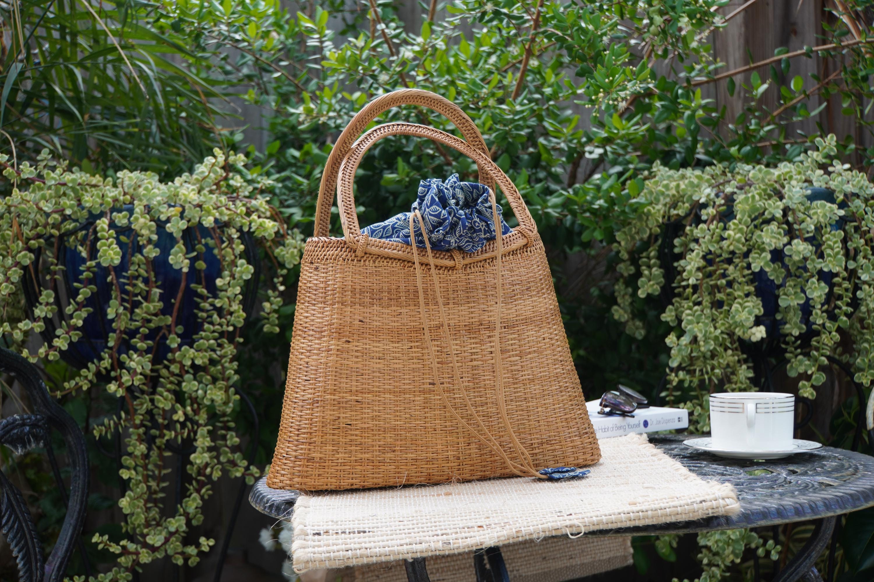 A woven straw handbag with a blue cloth tied around it is placed on a small table, with a white cup and a book nearby. The setting appears to be a garden or outdoor area with lush greenery in the background.