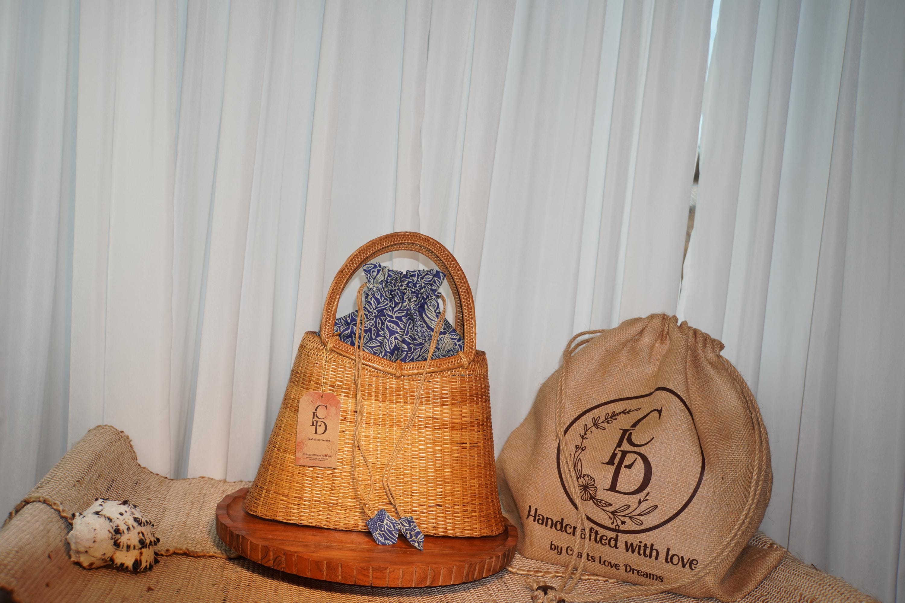 a woven straw bag with a blue and white patterned fabric, placed on a wooden surface next to a burlap sack with a logo.