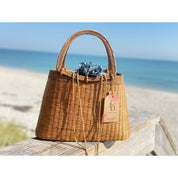 A brown woven handbag with a tag attached, placed on a wooden surface near the ocean.