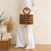 A person wearing white pants and holding a brown woven handbag stands in front of a beige wall with a wicker basket and some decorative pillows.