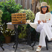 A woman in a white outfit is sitting on a bench, reading a book and holding a woven bag, with a potted plant and a small table nearby.