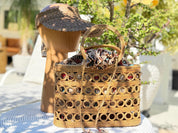 A woven straw bag with a patterned design sits on a table, accompanied by a wooden hat stand and a potted plant in the background.