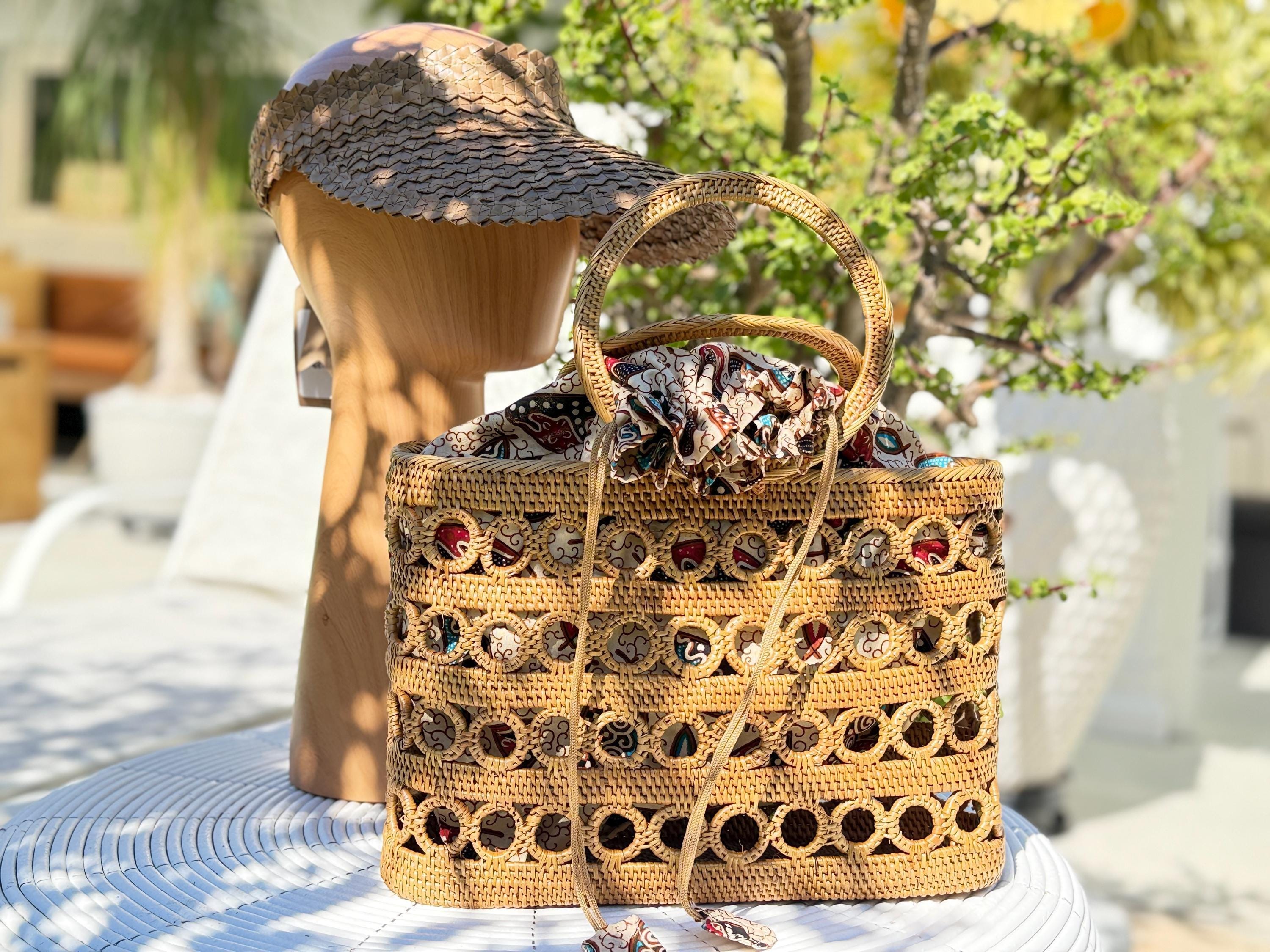 A woven straw bag with a patterned design sits on a table, accompanied by a wooden hat stand and a potted plant in the background.