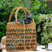 A woven straw bag with a pattern of triangles and a blue fabric draped over it, placed on a surface with greenery in the background.