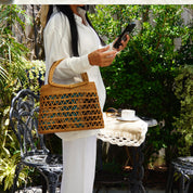 A woman in a white outfit stands in a garden, holding a woven purse and looking at her phone.