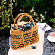 A woven straw handbag with a floral pattern sits on a white tablecloth, accompanied by a pair of glasses, a white napkin, and a small white bowl of tea.