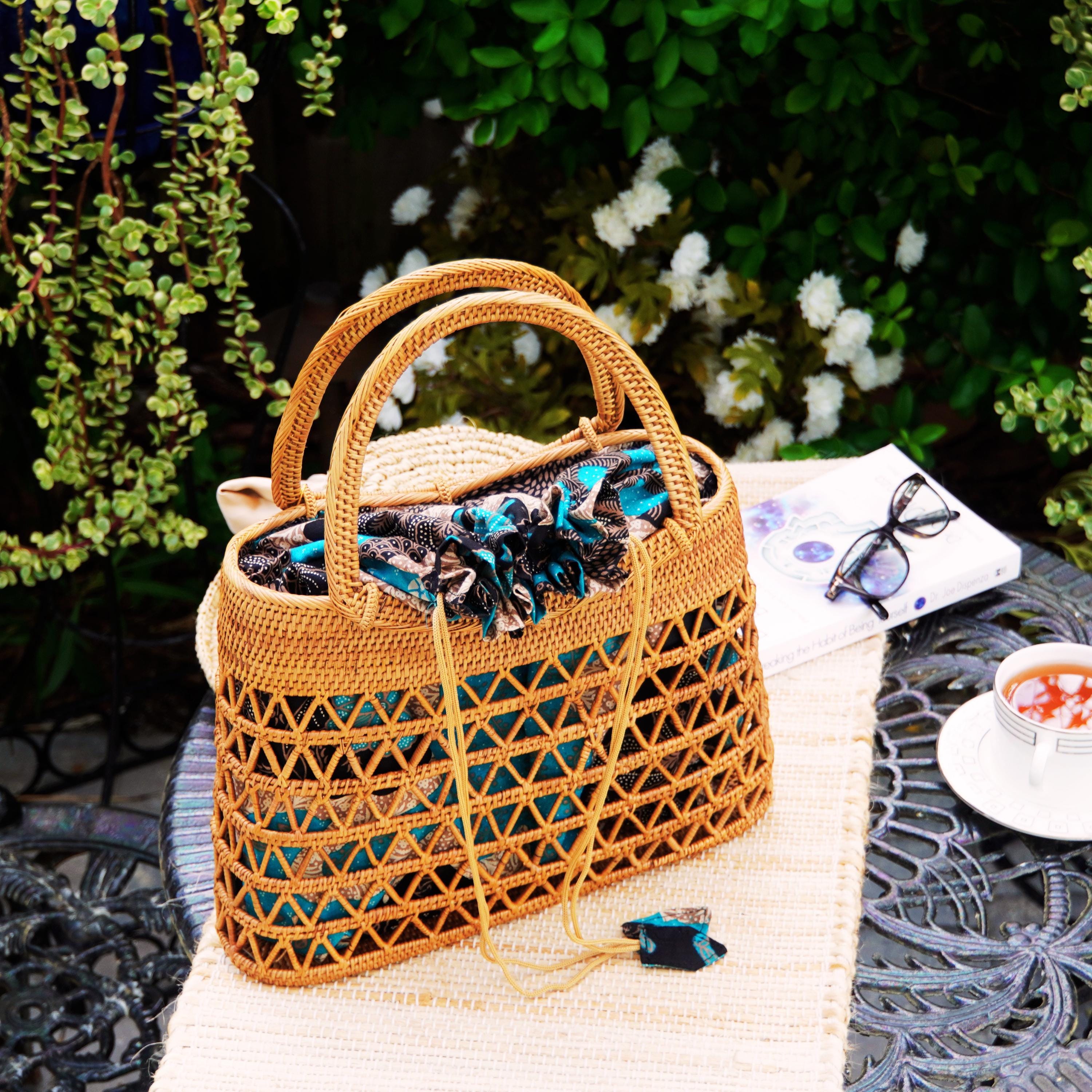 A woven straw handbag with a floral pattern sits on a white tablecloth, accompanied by a pair of glasses, a white napkin, and a small white bowl of tea.