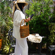 A woman in a white outfit is standing in a garden, holding a woven bag and looking at her phone.