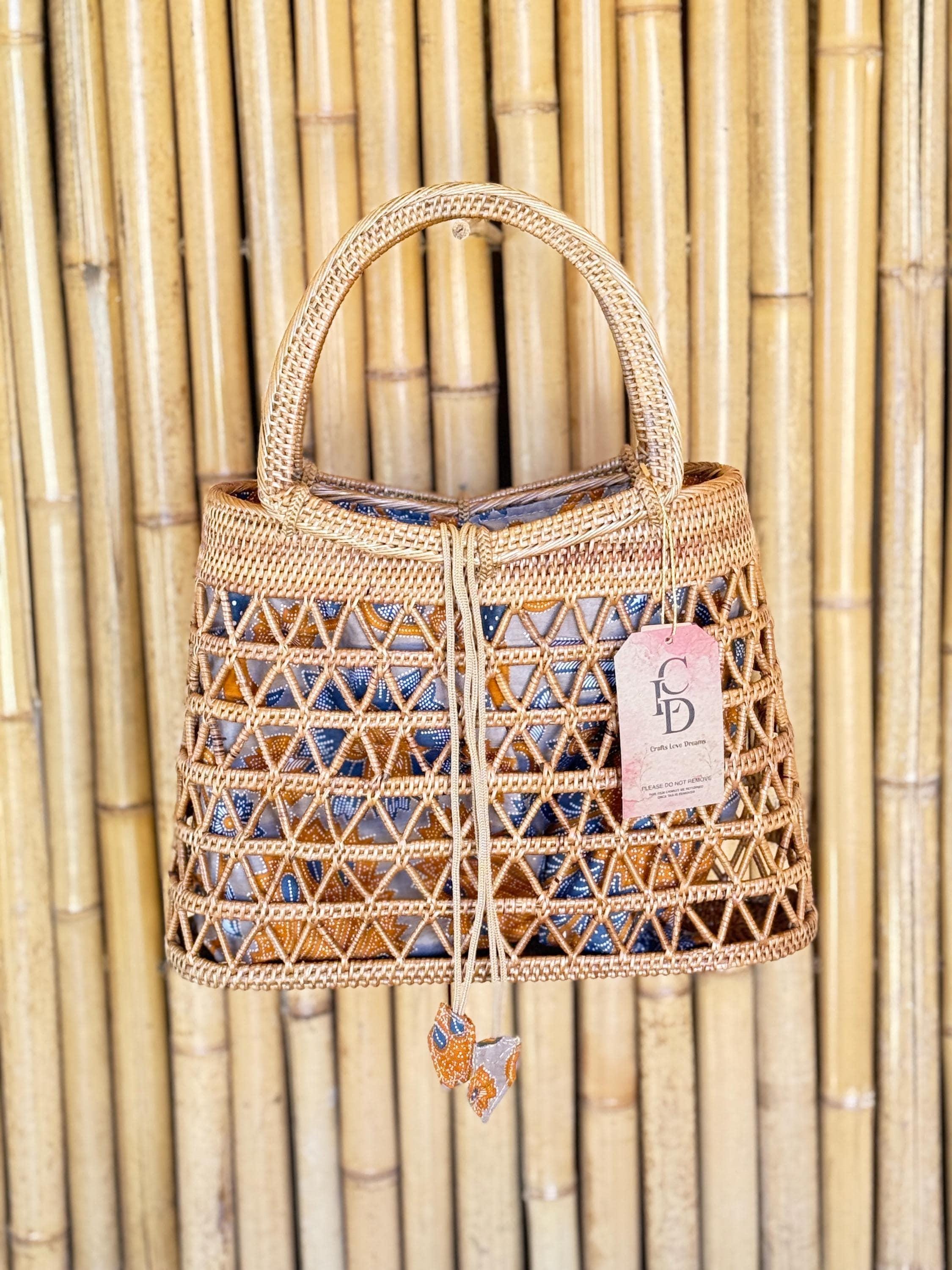 A woven handbag with a handle and a tag attached, displayed against a bamboo wall.