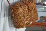 A brown woven purse with a leather strap is hanging on a white fabric background.
