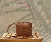 A brown woven purse with a leather strap is displayed on a wooden surface, with a blurred background that includes pink flowers and a screen.