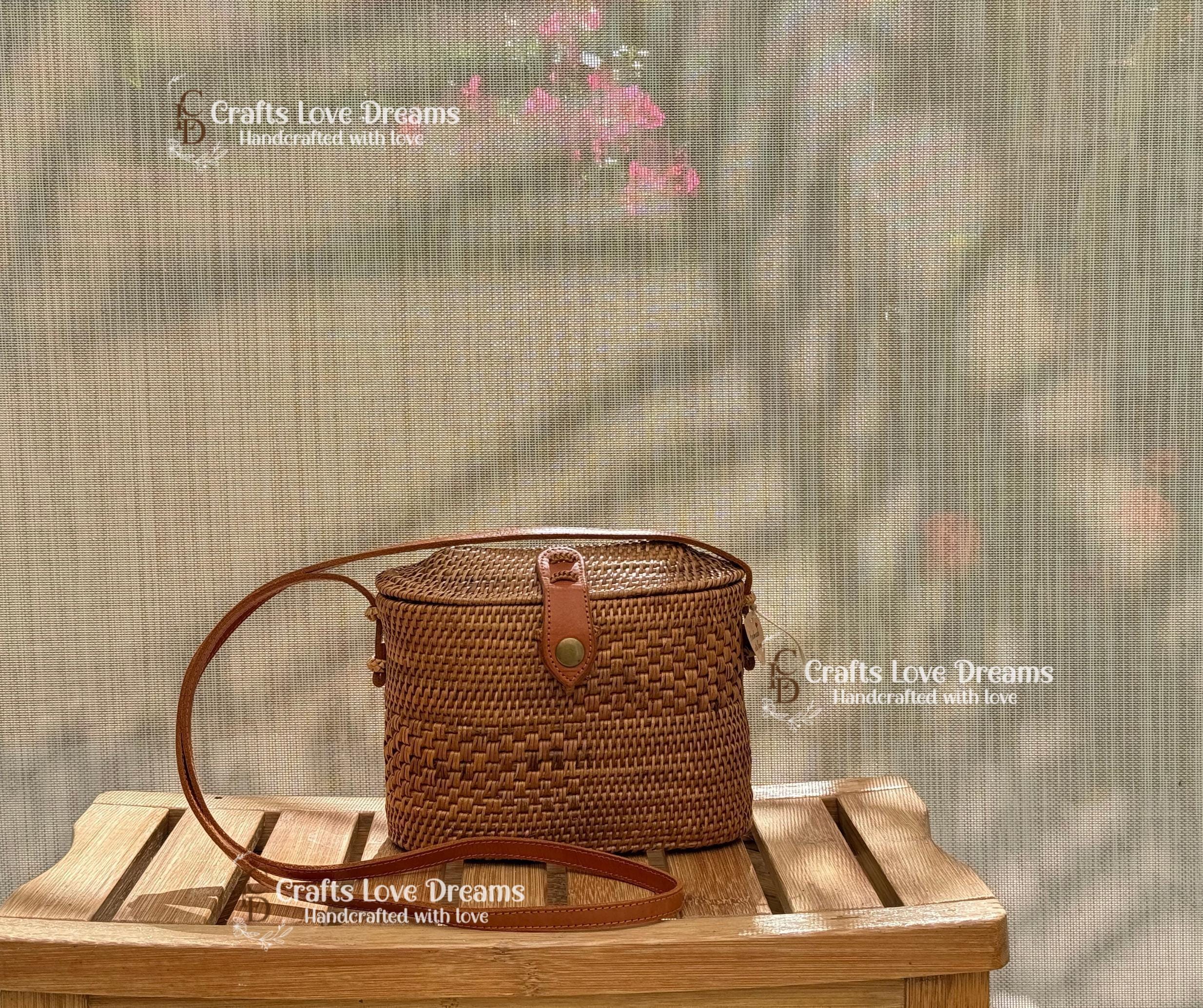 A brown woven purse with a leather strap is displayed on a wooden surface, with a blurred background that includes pink flowers and a screen.