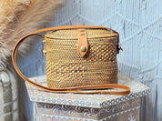 A woven straw bag with a leather strap is displayed on a decorative wooden stand against a white wall.
