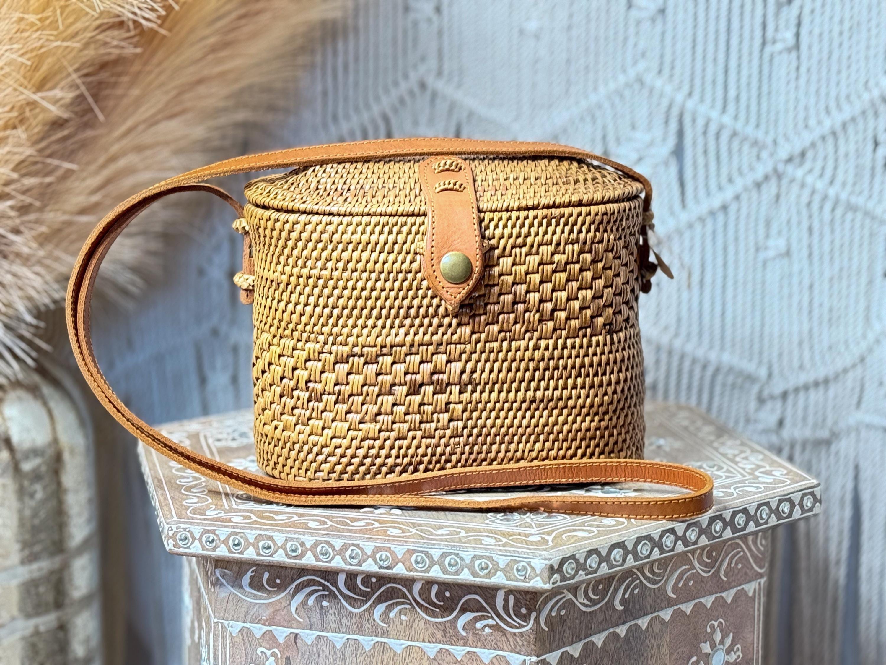 A woven straw bag with a leather strap is displayed on a decorative wooden stand against a white wall.