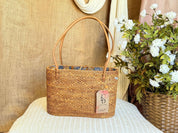 A woven straw handbag with a tag attached, placed on a white surface next to a potted plant with white flowers.