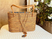 A woven straw bag with a brown leather handle is displayed on a white surface, with a vase of white flowers in the background.