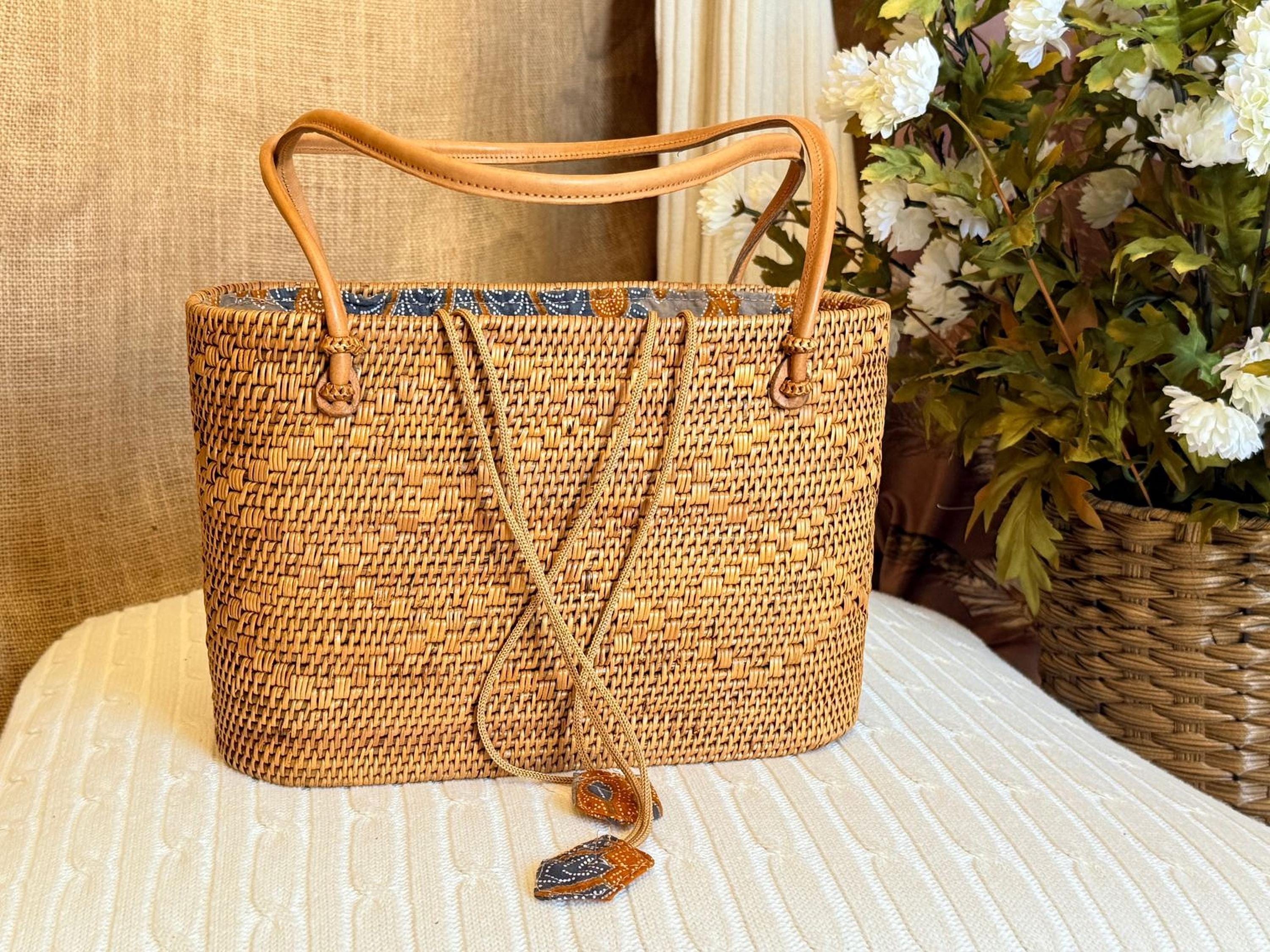 A woven straw bag with a brown leather handle is displayed on a white surface, with a vase of white flowers in the background.