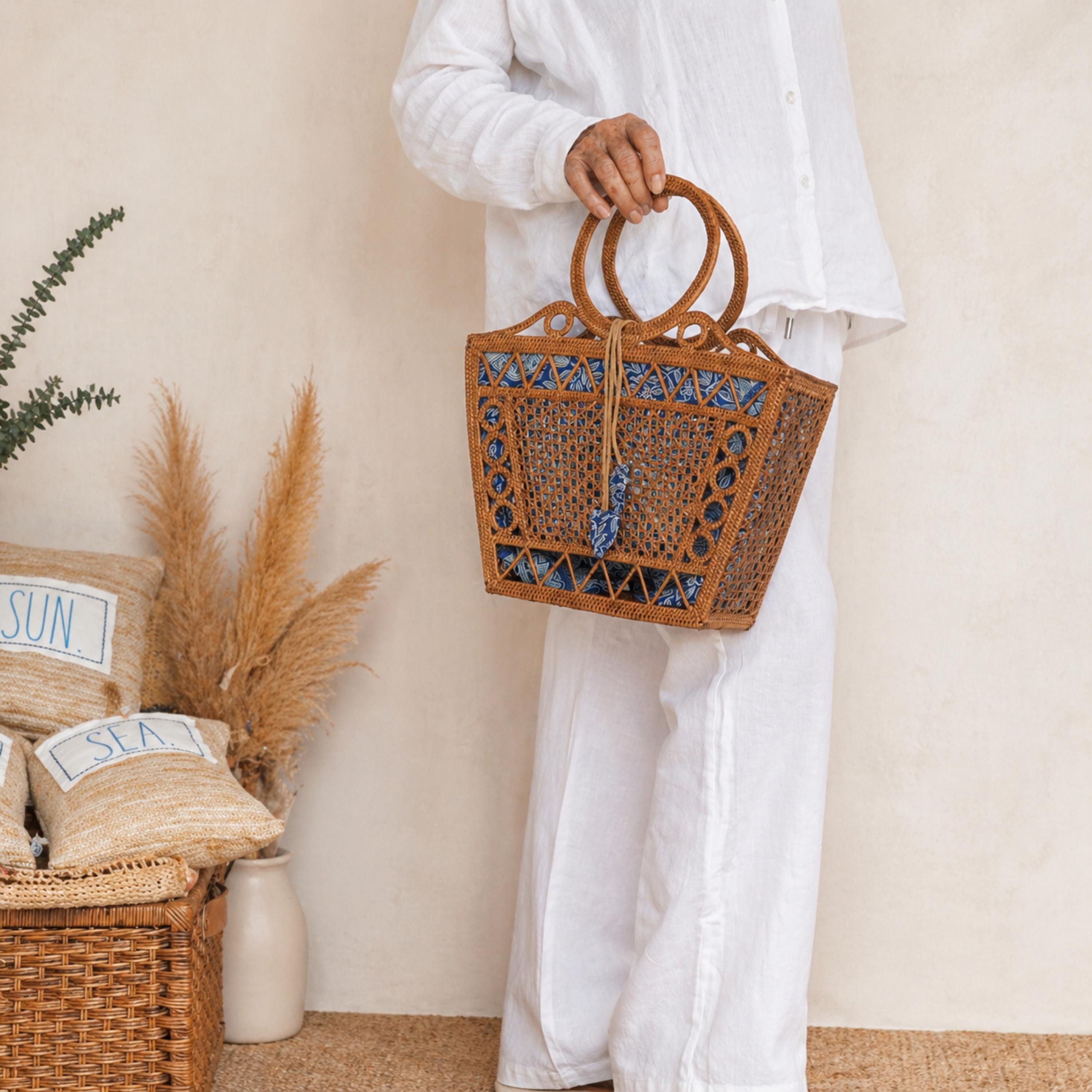 A person wearing white pants and holding a brown woven handbag stands in front of a beige wall with a potted plant and a woven basket.