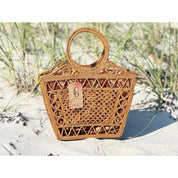 A woven straw handbag with a round handle and a tag attached, placed on a sandy beach with some grass in the background.