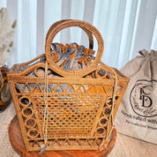 A woven handbag with a round handle and a drawstring closure sits on a wooden surface, accompanied by a burlap sack with the text "Handcrafted with love dream" printed on it.