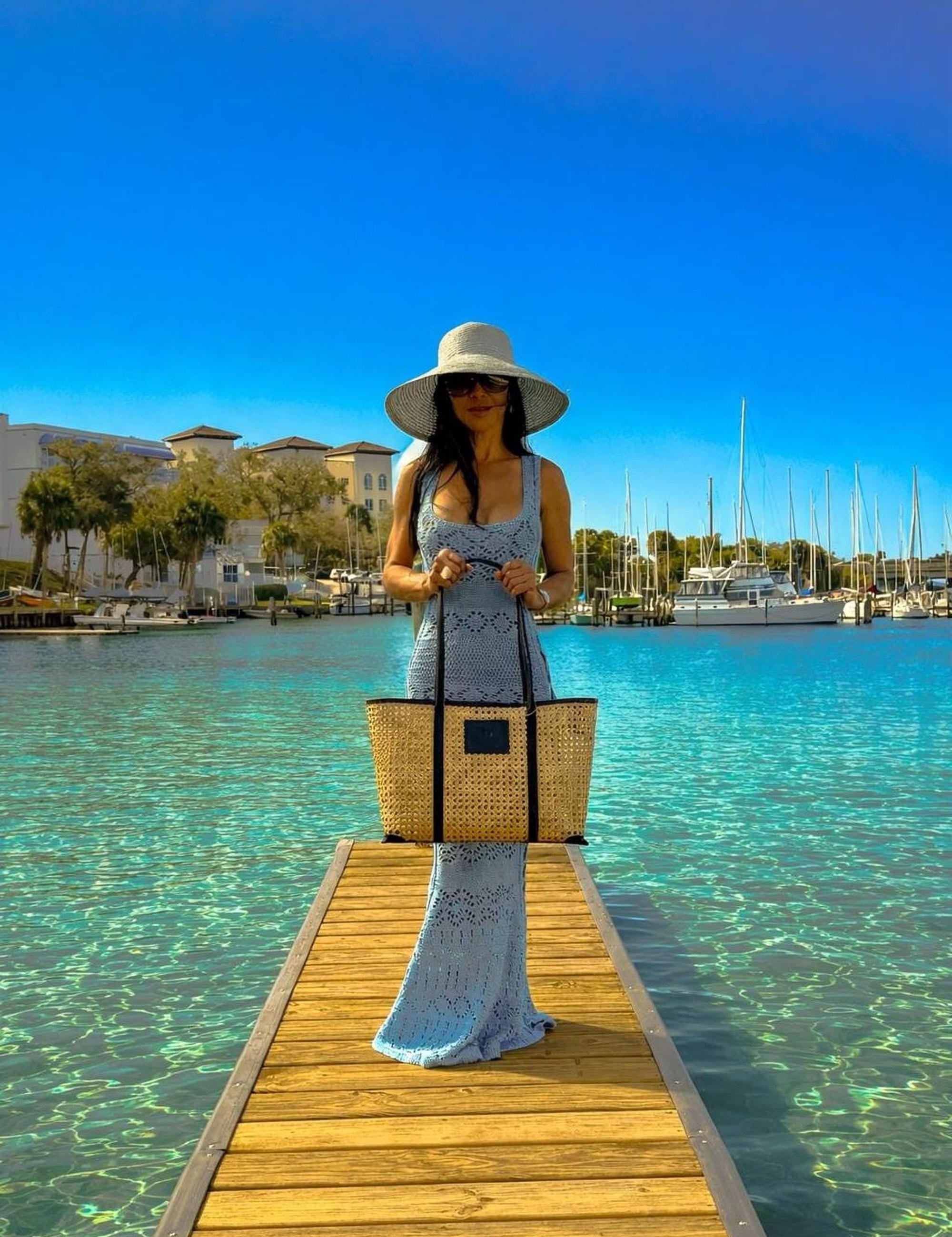 A woman in a blue dress and wide-brimmed hat stands on a wooden pier, holding a large woven bag, with a marina and sailboats in the background.