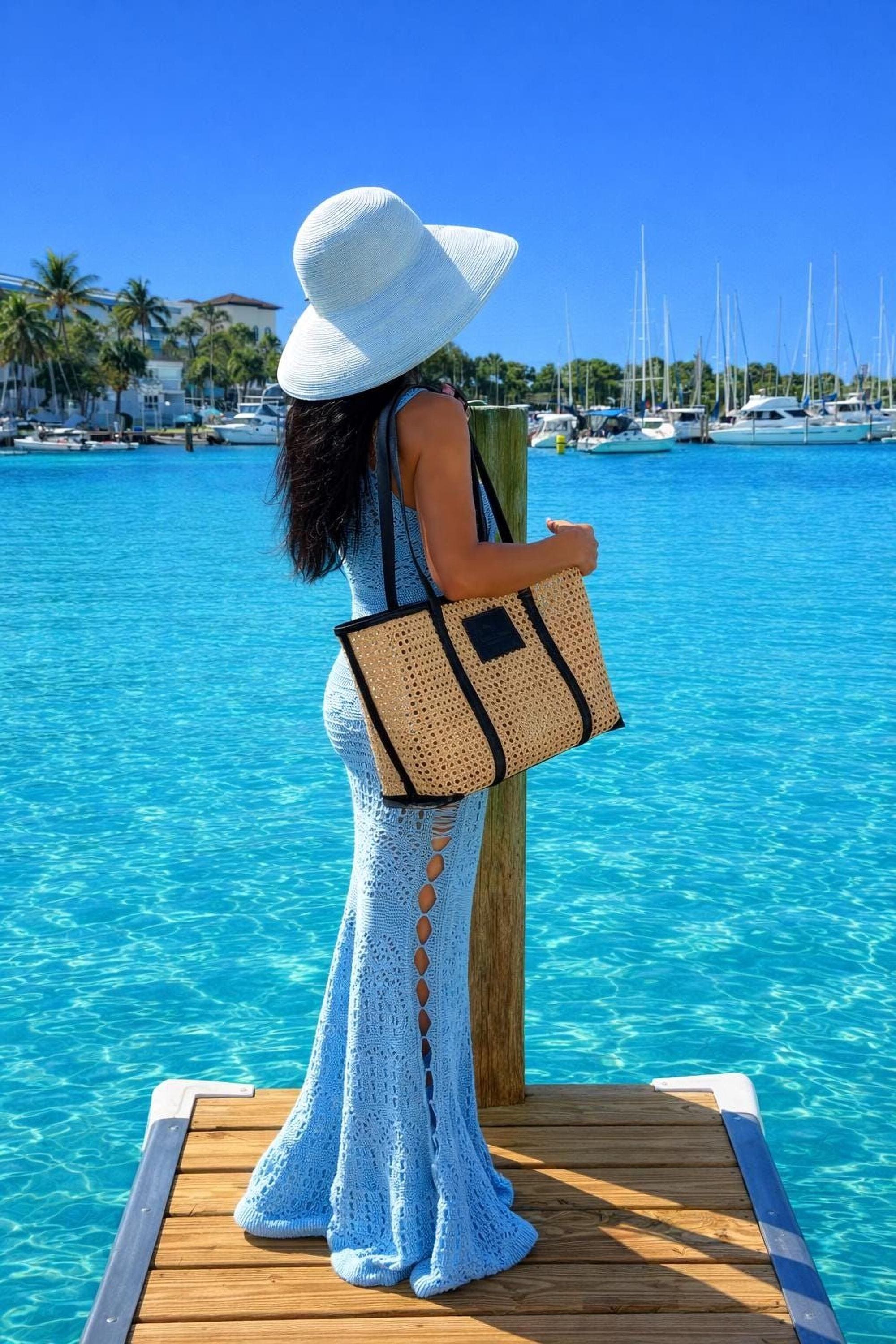A woman in a blue dress and white hat stands on a wooden dock, gazing out at a marina filled with sailboats and yachts.