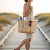 A woman is walking on a wooden boardwalk, carrying a large woven tote bag with a blue and white striped towel inside.